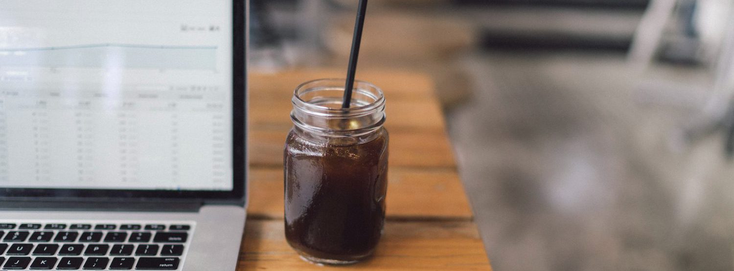 Iced coffee in a mason jar with a black straw next to a laptop computer.