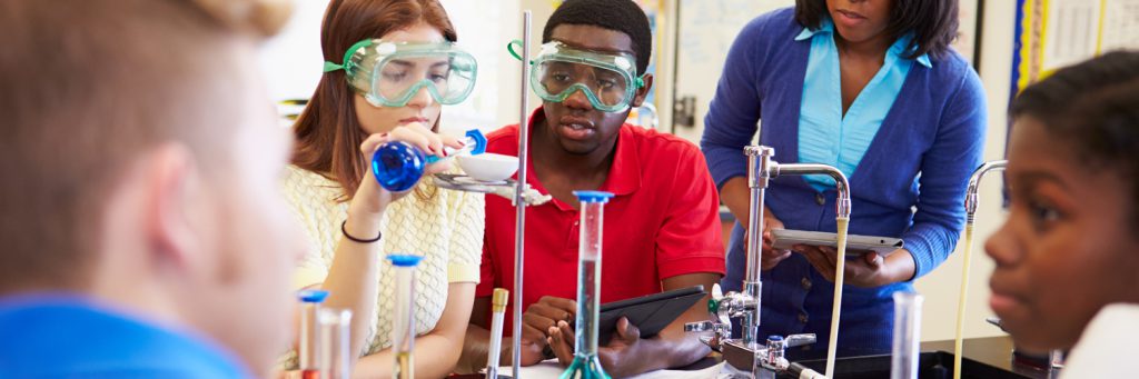 two students doing a science experiment following lab safety rules with the teacher watching close by